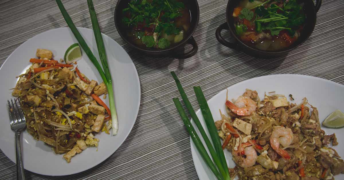 Stojące na stole dwie miseczki z zupą Tom Yum oraz dwa talerze z Pad Thai