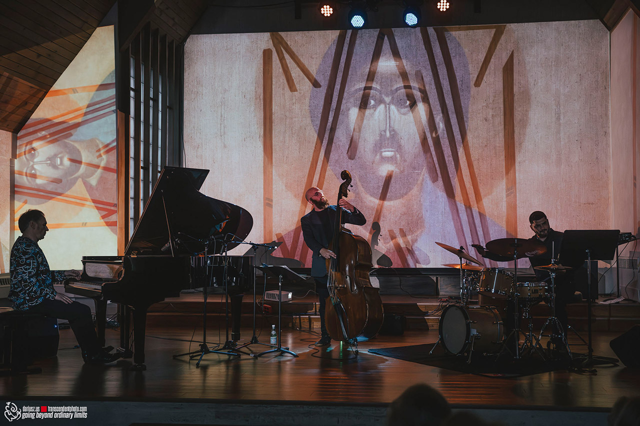 Artur Dutkiewicz Trio podczas koncertu na scenie; w tle na dużym ekranie widoczna jest nowoczesna, podświetlona na czerwono ikona przedstawiająca twarz Jezusa Chrystusa.