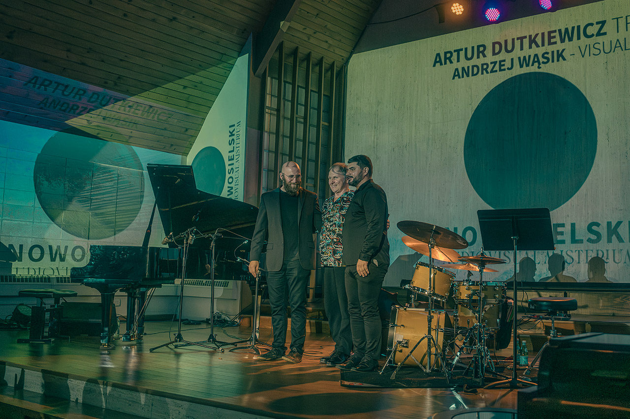 Trzej muzycy jazzowi z Artur Dutkiewicz Trio stojący na scenie obok fortepianu i perkusji, na tle nowoczesnej scenografii z okrągłymi grafikami w Akademii Muzyki PaSO.