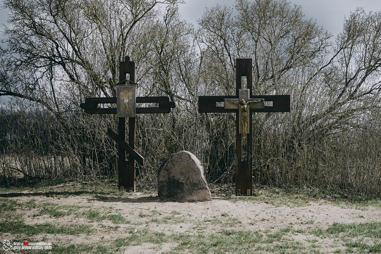 Tradycyjny krzyż prawosławny oraz krzyż katolicki stojące obok siebie na otwartej przestrzeni na Podlasiu, z pamiątkowym kamieniem u podstawy i lasem w tle.