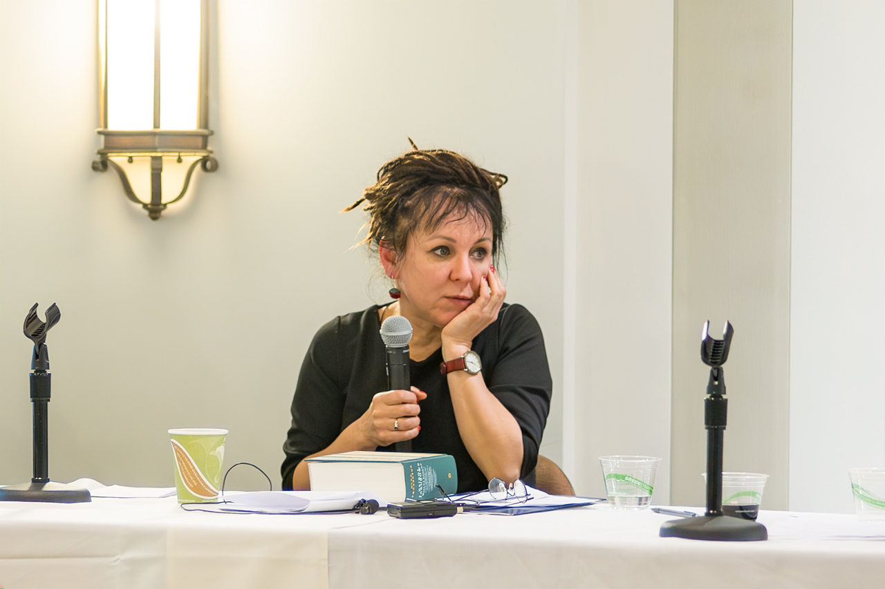 Pisarka Olga Tokarczuk siedząca przy stole z mikrofonem i książką podczas spotkania autorskiego w Chicago, zamyślona, z dłonią opartą na podbródku.