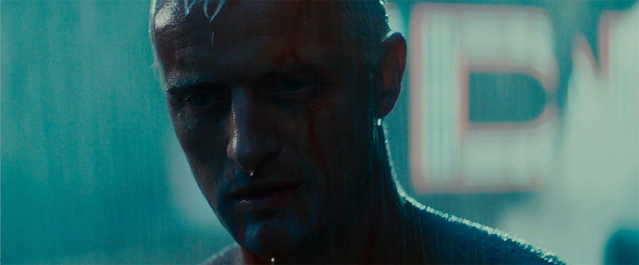 Bliski kadr na twarz Rutgera Hauera jako replikanta Roya Batty'ego z filmu „Blade Runner”, zalaną deszczem, ze spływającymi po niej kroplami wody i łzami, w neonowym oświetleniu futurystycznego miasta.