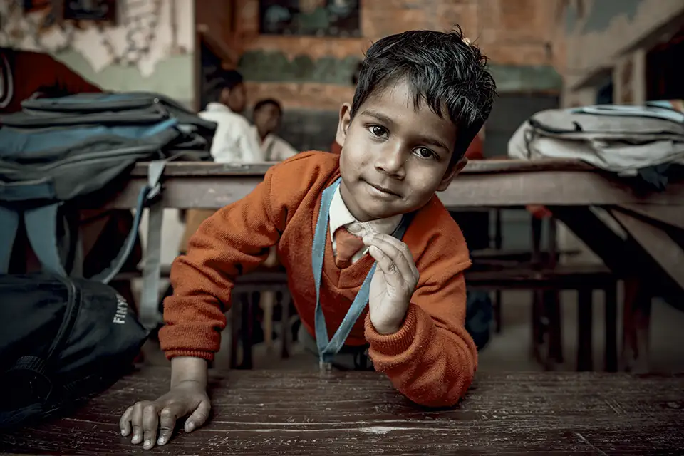 Young boy in orange sweater looks curiously at camera in classroom.