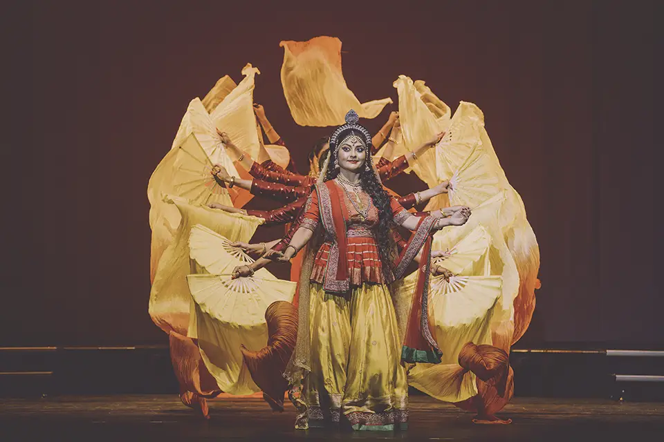 Indian dancer portraying goddess with multiple arms and yellow fans.
