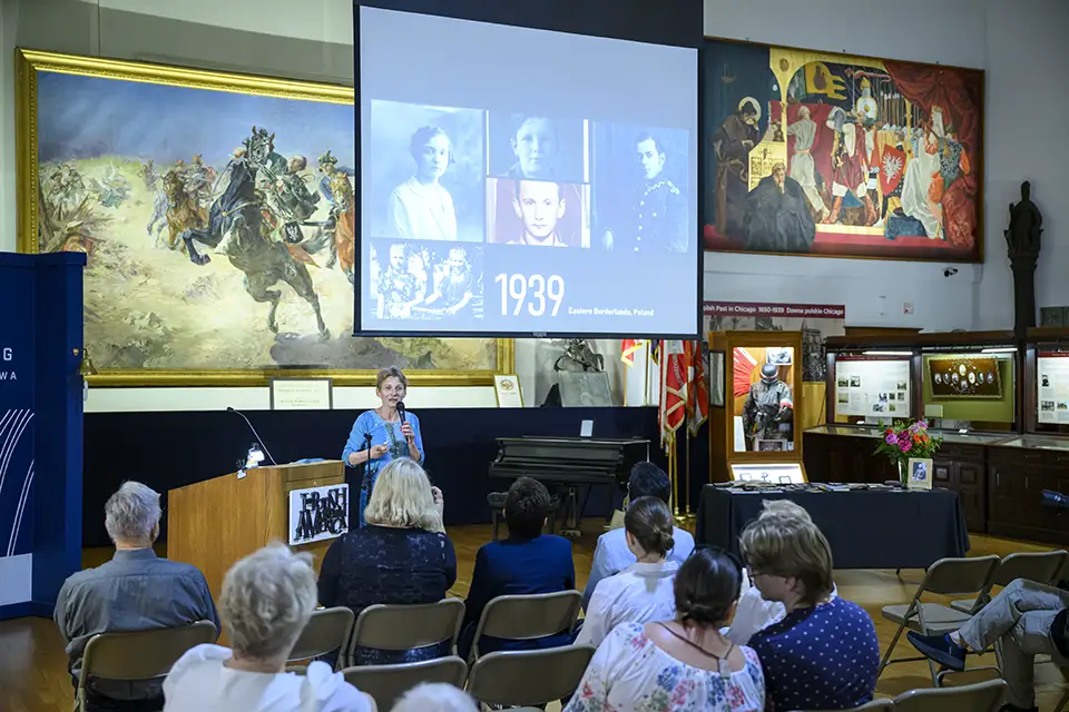 Woman lecturing to audience in museum with historical paintings.