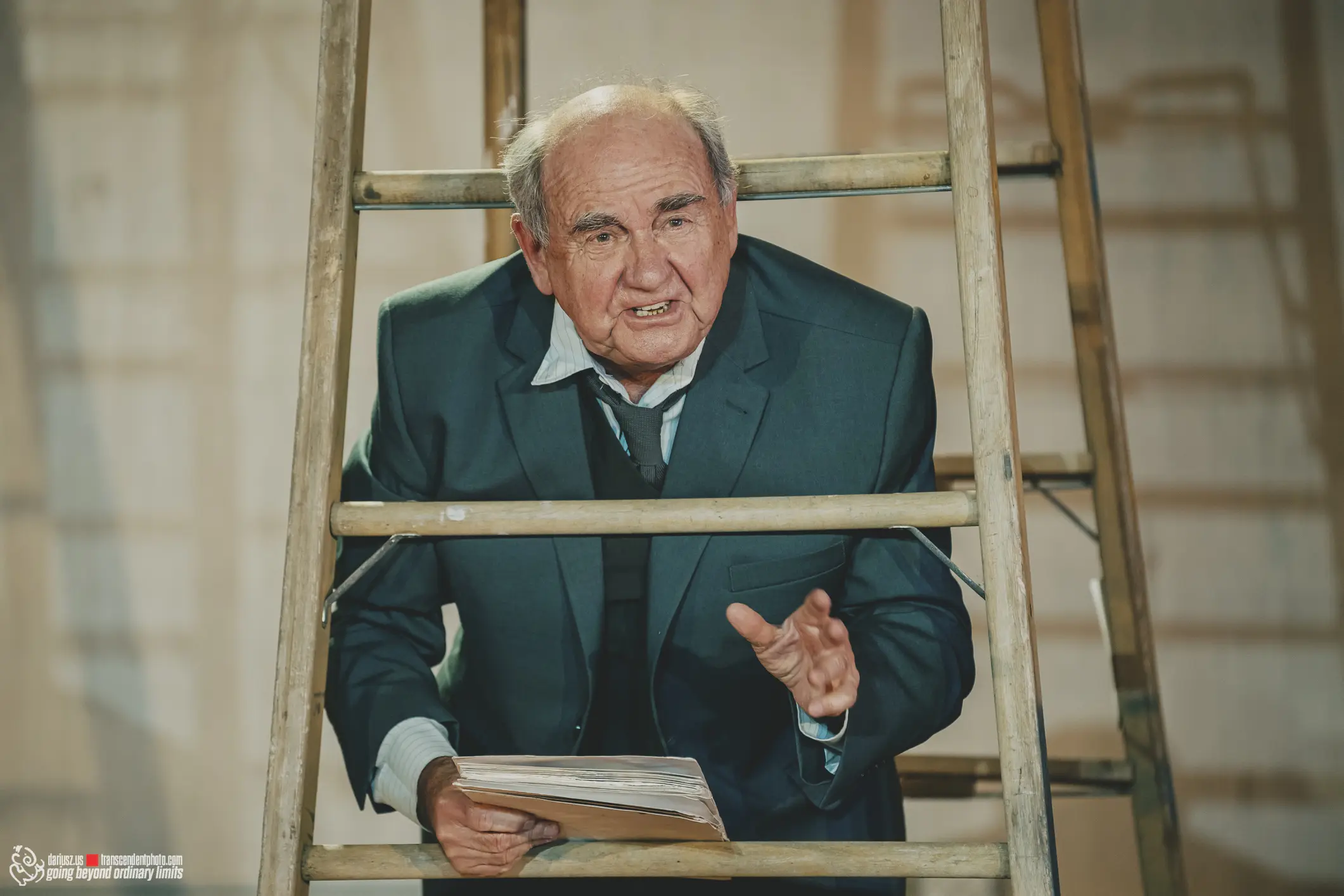 Older man in suit behind ladder, holding book, expressive face.