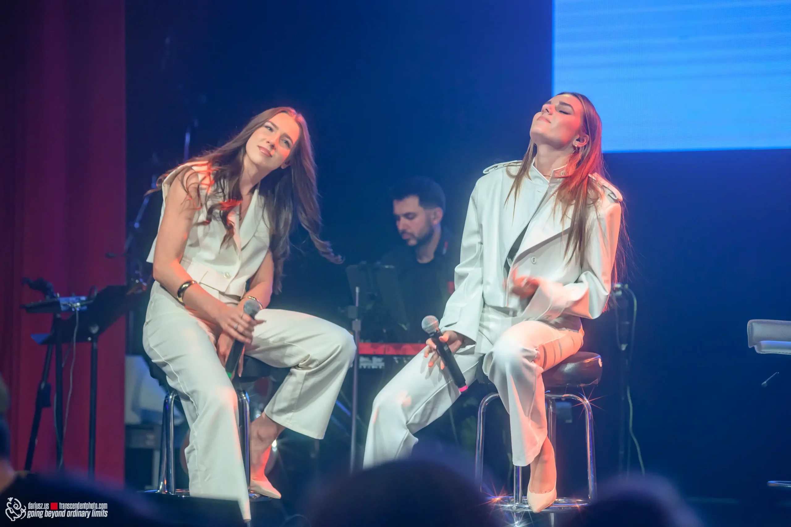 Two women performing on stage in white outfits, emotional expressions.