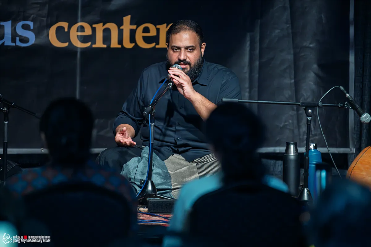 Bearded man speaking into microphone on stage to an audience.