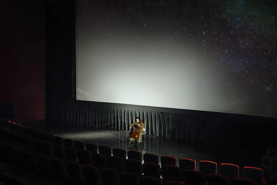 Cellist performs on stage in empty cinema with starry screen.