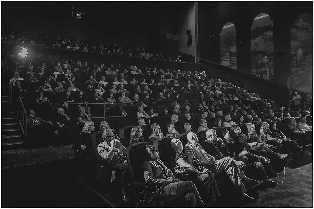 Audience in a dimly lit theater watching a performance in black and white.