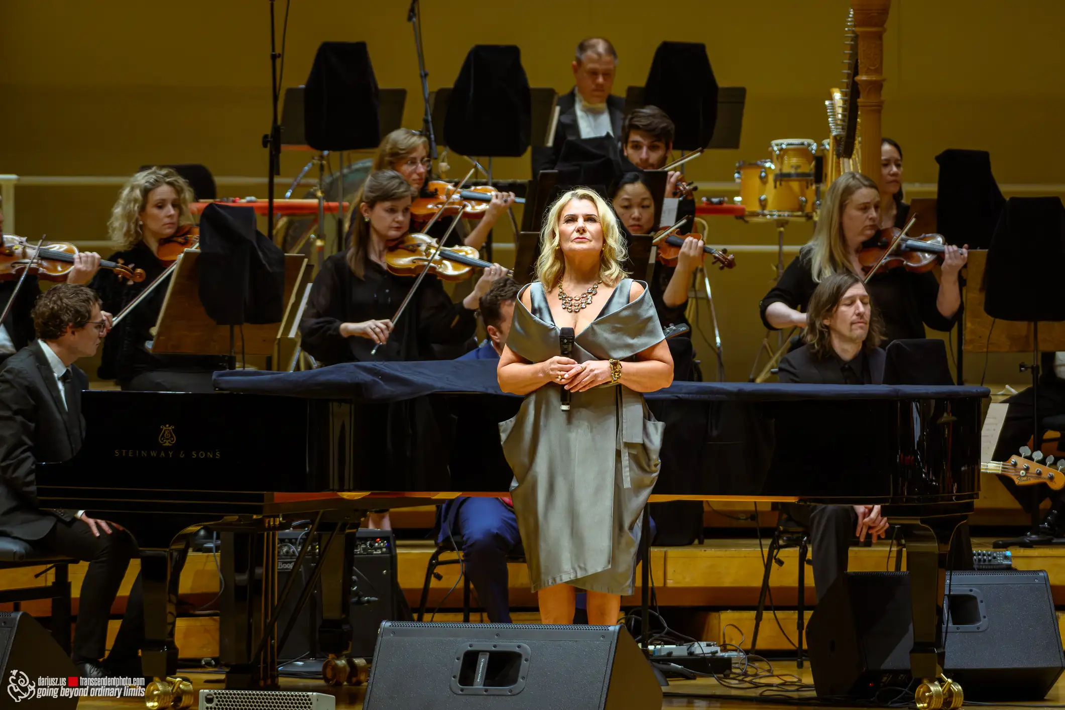 Blonde singer performs with orchestra and grand piano on stage.