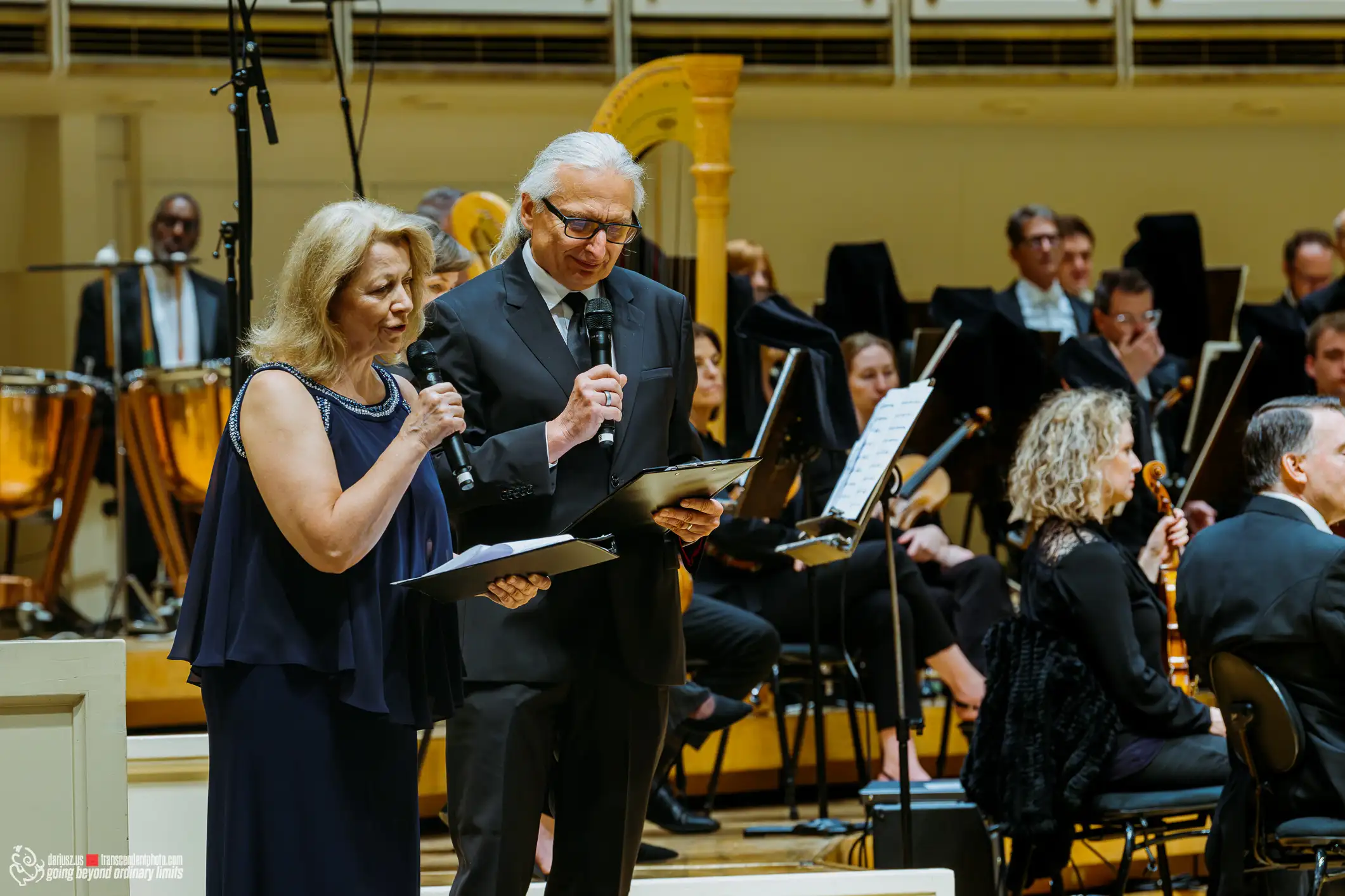 Presenters speak to an orchestra audience, with musicians and instruments visible.