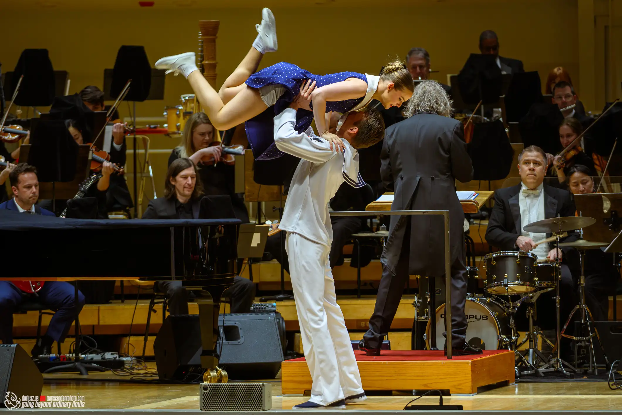 Sailor dancer lifts partner mid-air during performance with orchestra.